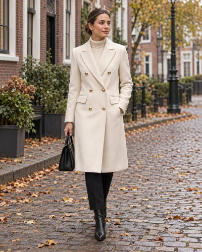 Vrouw in een knielange beige winterjas met gouden knopen en een zwarte tas op een keienstraat in de herfst