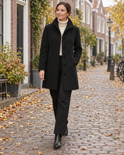 Vrouw in een klassieke halflange zwarte jas met witte col wandelt over een straat vol herfstbladeren