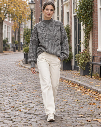Vrouw in een grijze grofgebreide trui met opstaande kraag en een crème broek in een Nederlandse straat